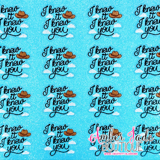 A repeating pattern of the phrase 'I knew it I knew you' in black script, each cowboy hat illustrated in brown with a white cloud behind it, all on a bright blue glitter-textured background. A 'Amber Jade Boutique' watermark is visible in the lower right corner.